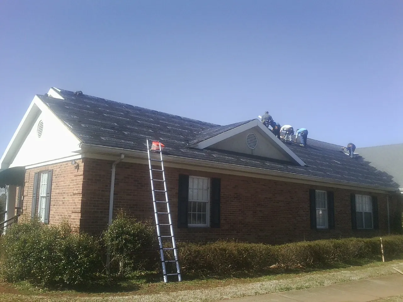 Crew tearing shingles off a brick ranch roof with a ladder set — Emergency Roof Repair work by our crew in Mendon